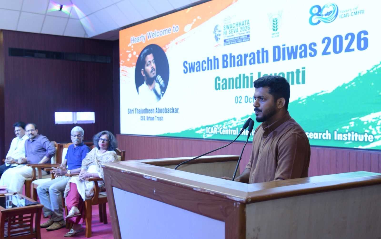 A man in a brown shirt speaking at a lectern with two microphones, with four guests seated beside the stage and a screen behind reading Swachh Bharath Diwas 2026, Gandhi Jayanti.