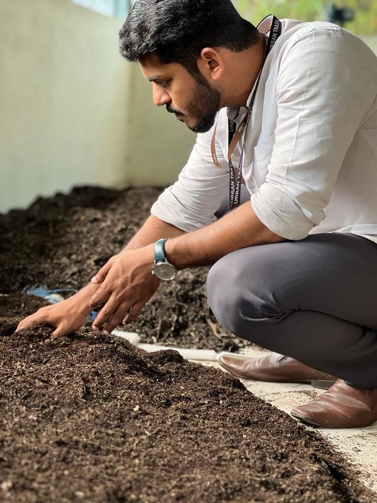 A man in a white shirt crouching beside a long bed of dark compost, working it with his hands.