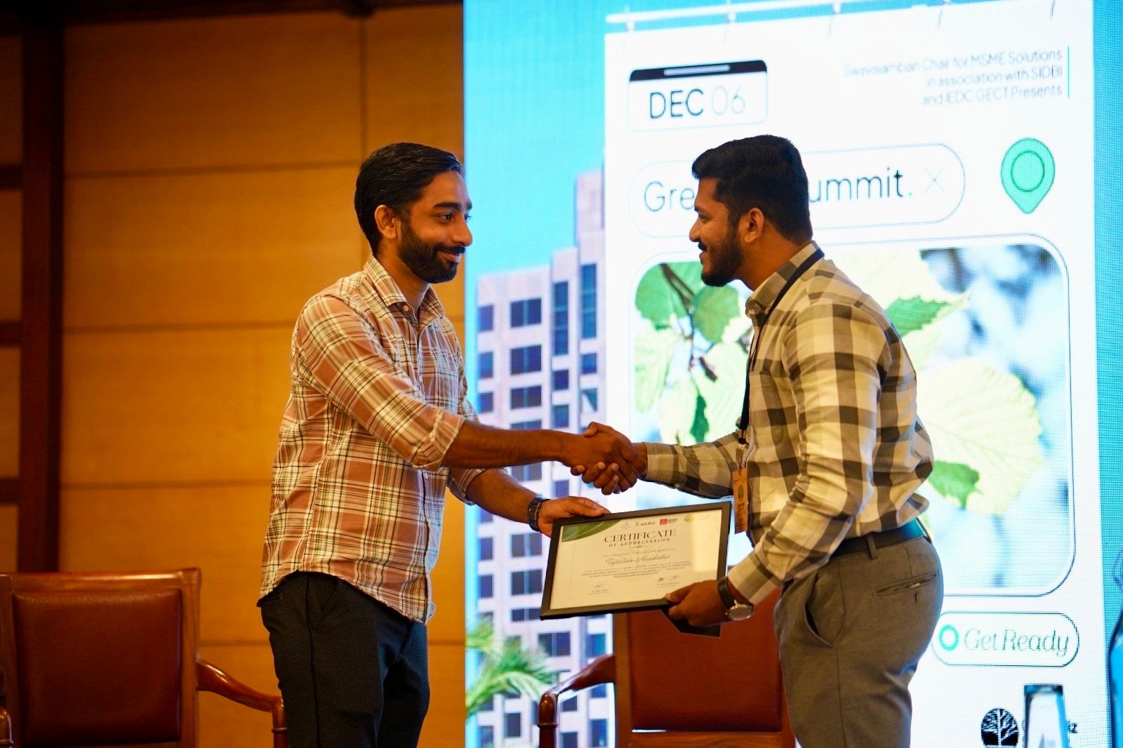 Two men shaking hands on stage, one holding a framed certificate, with a Green Summit slide behind them.