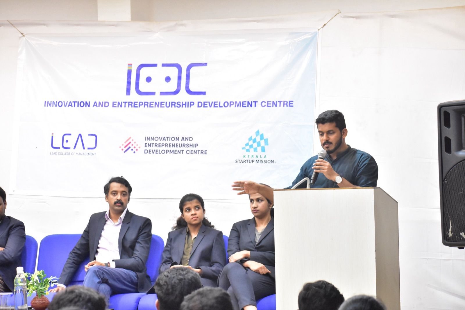 A man in a dark shirt speaking from a lectern, with three seated panellists and an Innovation and Entrepreneurship Development Centre banner behind.