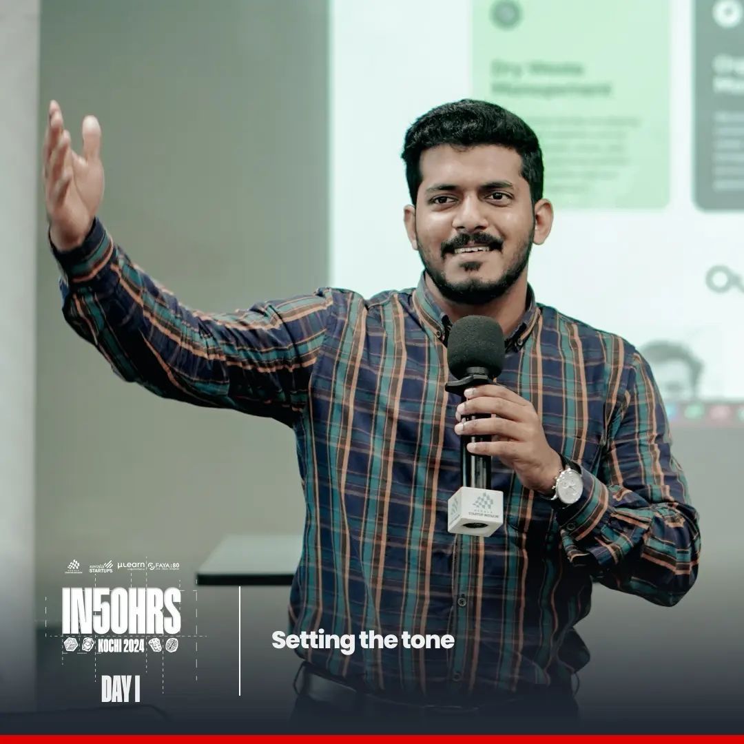 A man in a checked shirt holding a microphone and gesturing with one hand, with a presentation slide behind him.