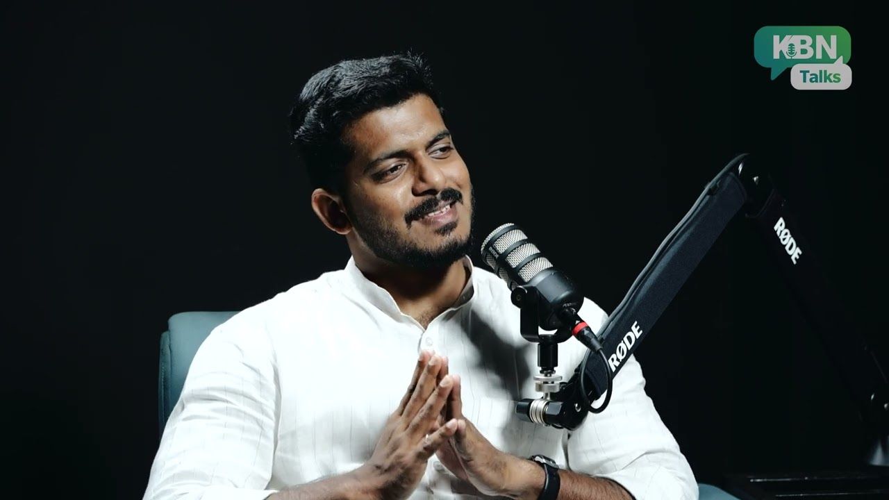 A man in a white shirt with his hands together, speaking in a podcast studio with a microphone on a boom arm.