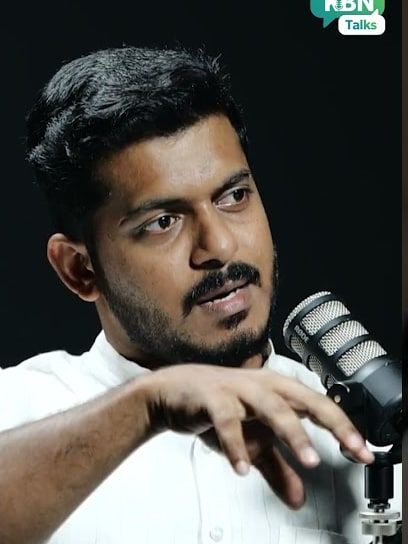 A man in a white shirt speaking into a studio microphone and gesturing with one hand.
