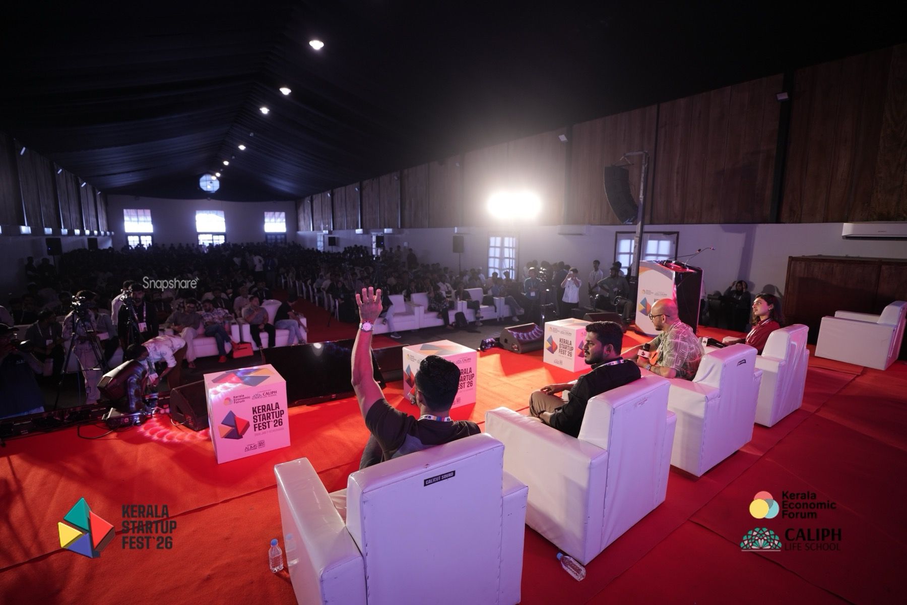 The view from the panel seats at Kerala Startup Fest 2026: a packed hall ahead, cameras along the edge, and one panelist with a raised hand.