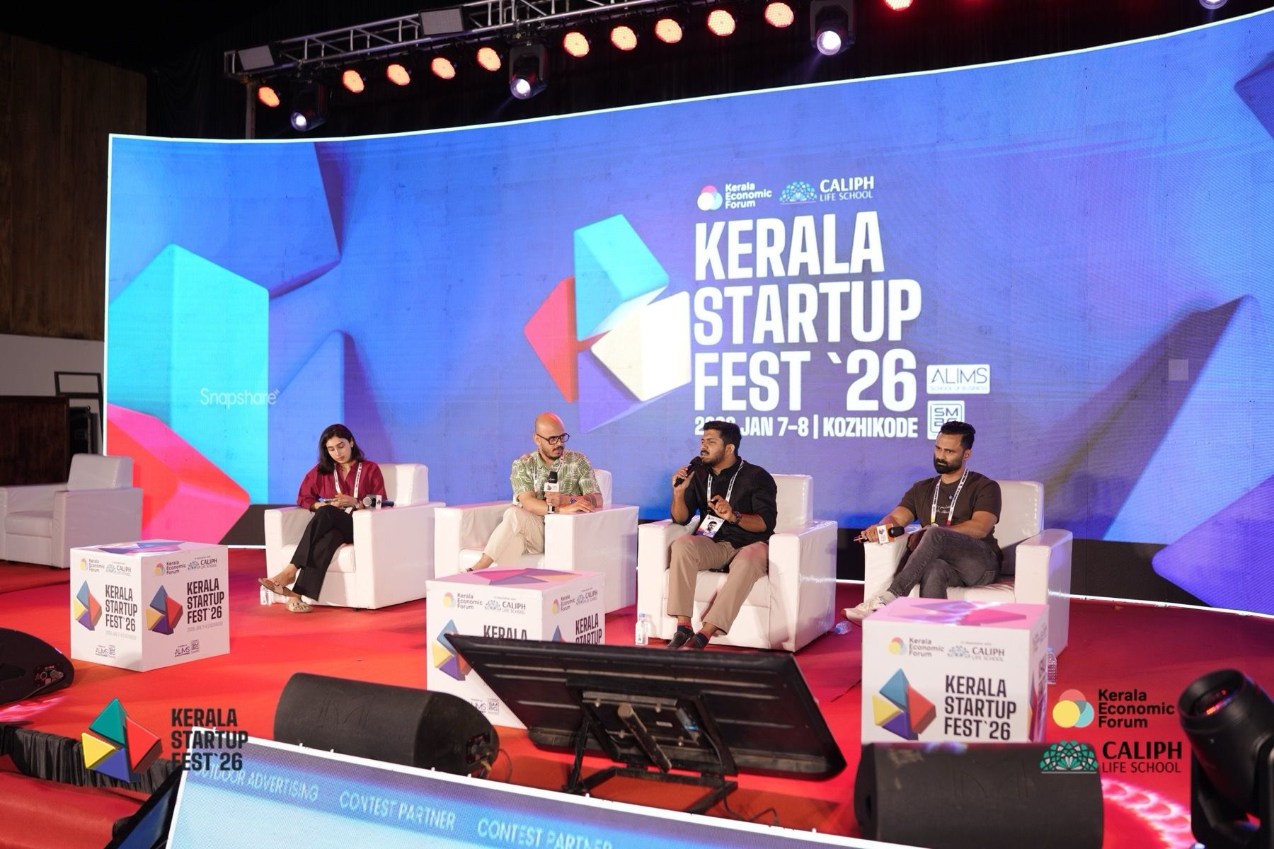 Four people on a panel sit in white armchairs on a red-carpeted stage, in front of a large screen that reads Kerala Startup Fest 2026, Kozhikode. The second from the right is speaking.