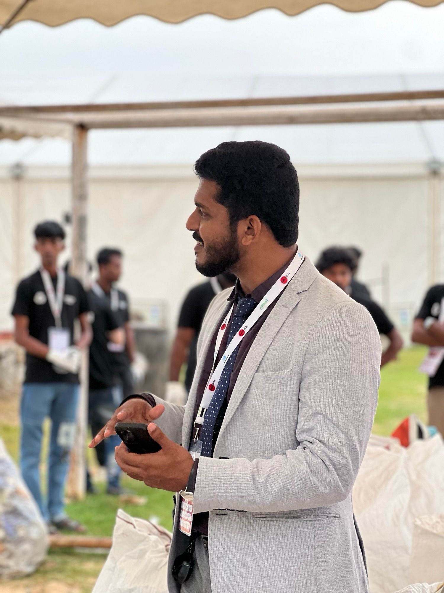 A smiling man in a grey jacket and lanyard holds a phone, with team members in black shirts out of focus behind him.