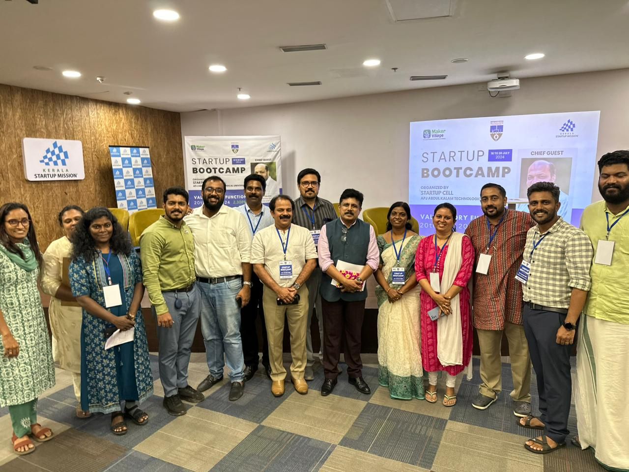 About fifteen people stand shoulder to shoulder in a hall, in front of a screen and banner for the Startup Bootcamp, 18 to 20 July 2024.