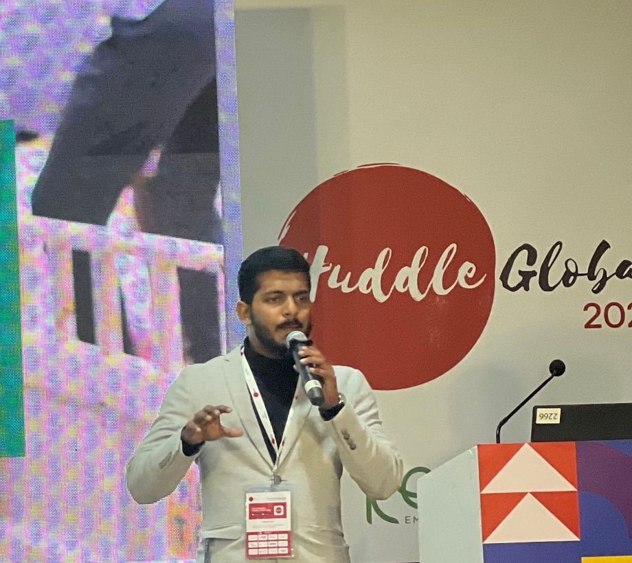 A man in a light grey jacket and lanyard speaks into a microphone beside a lectern, in front of a Huddle Global backdrop.