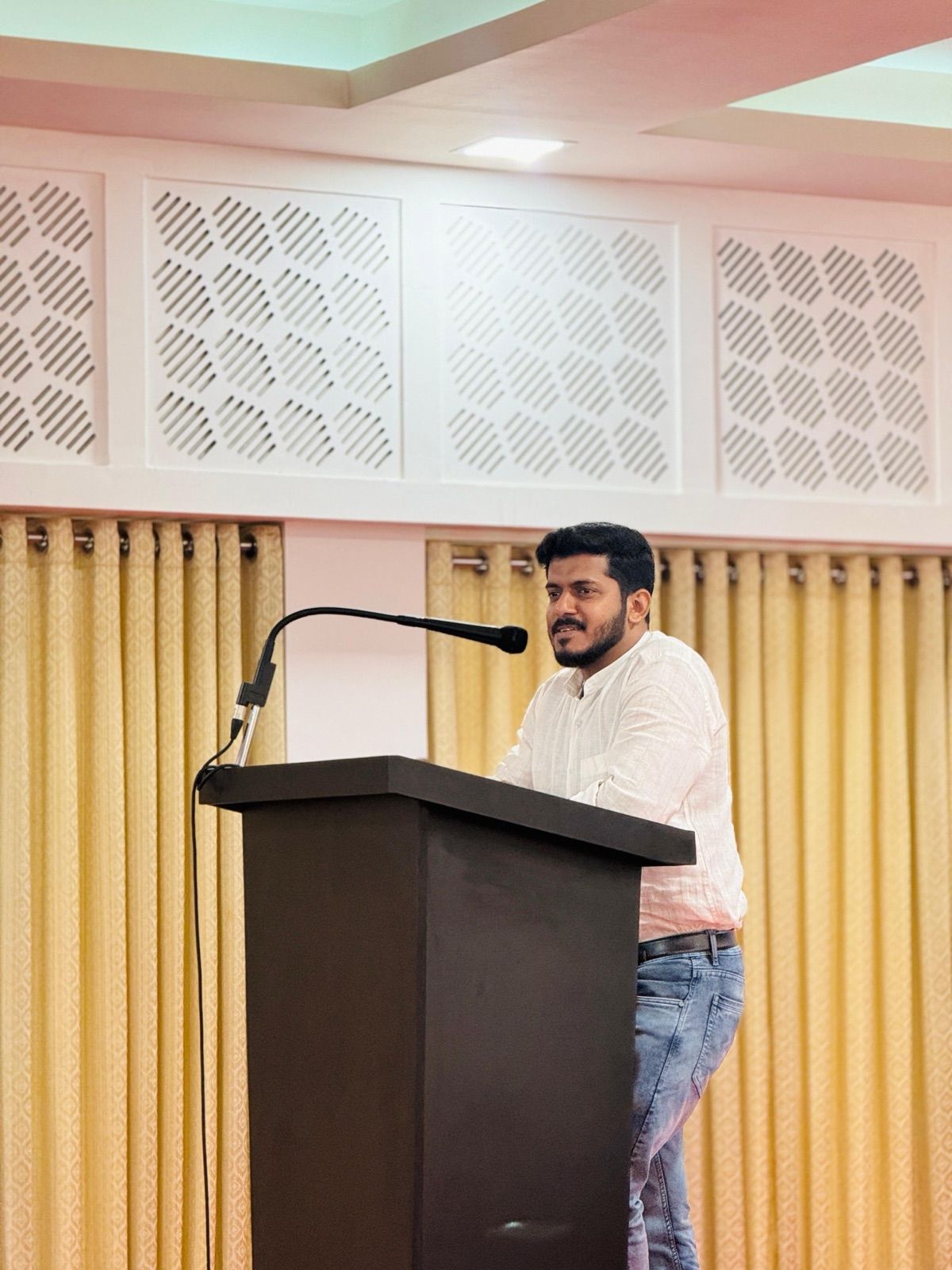 A man in a white shirt speaking at a dark lectern with a microphone, in a hall with gold curtains.