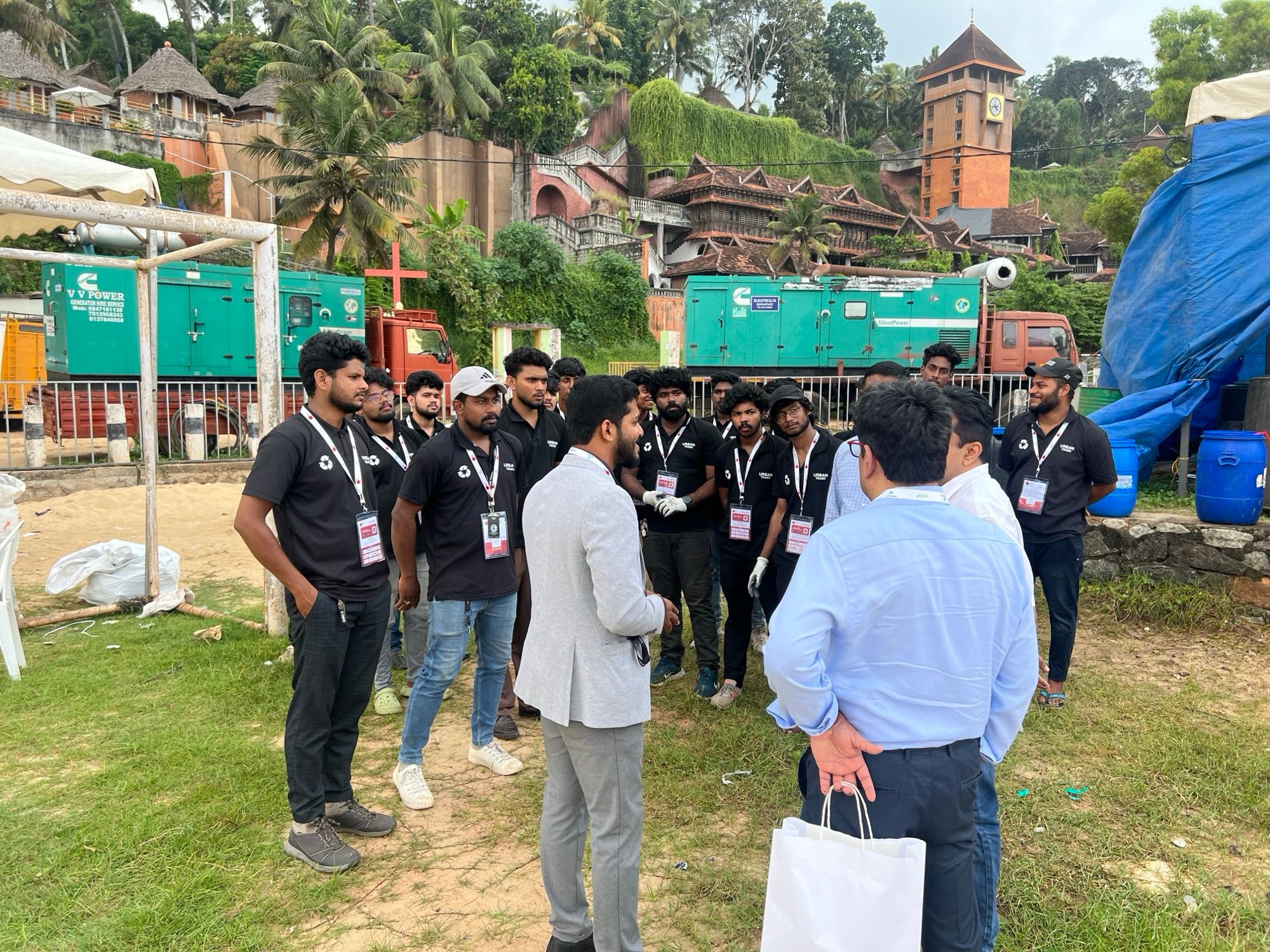 A man in a light grey jacket speaking to a group of team members in black polo shirts on a grass field, with green generator trucks behind them.